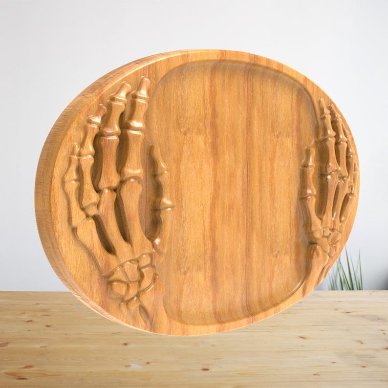K&ouml;nnte beinhalten: Ein rundes Holztablett mit einem geschnitzten Relief von zwei Skeletth&auml;nden auf der linken und rechten Seite. Das Tablett hat eine nat&uuml;rliche hellbraune Holzmaserung mit einer glatten, vertieften Mitte zum Ablegen von Gegenst&auml;nden.