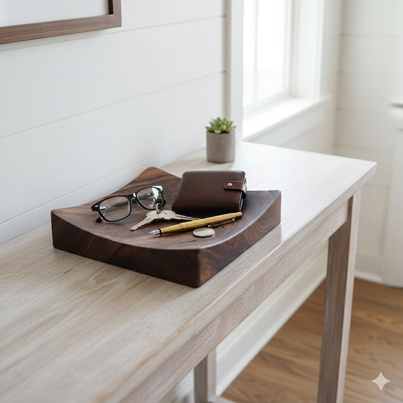 K&ouml;nnte beinhalten: Ein dunkelbraunes Holztablett mit geschwungenem Design enth&auml;lt Brille, Schl&uuml;ssel, eine braune Ledergeldb&ouml;rse, einen goldenen Stift und eine M&uuml;nze. Eine kleine Topfpflanze steht daneben. Das Tablett steht auf einem hellen Holztisch.