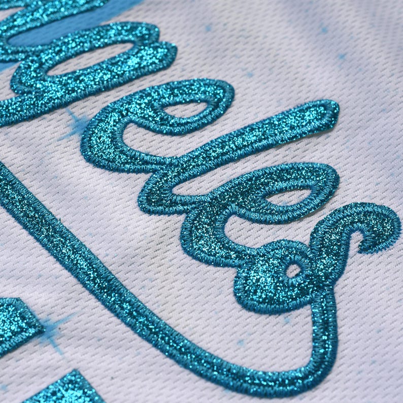 May include: Close-up of a white fabric with a teal glitter embroidered word. The teal embroidery has a sparkly texture. The background has a subtle blue star pattern.