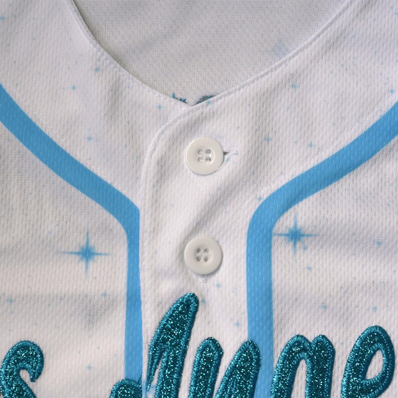 May include: White baseball jersey with light blue accents and sparkling embellishments. Features a V-neck, button-up front, and a design with a celestial theme. The jersey has a mesh-like texture and a decorative design.