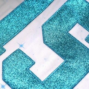 May include: Close-up of a white fabric with turquoise glitter-covered geometric shapes. The shapes are outlined with a white border. Small, light blue starbursts are scattered across the background.