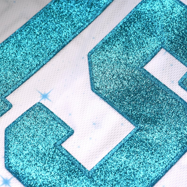 May include: Close-up of a white fabric with turquoise glitter-covered geometric shapes. The shapes are outlined with a white border. Small, light blue starbursts are scattered across the background.
