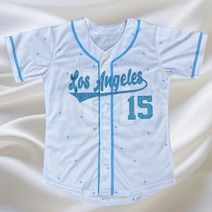 May include: White baseball jersey with light blue accents and the text "Los Angeles" in glittery blue script. The number "15" is also in glittery blue. The jersey has a button-up front and a V-neck.