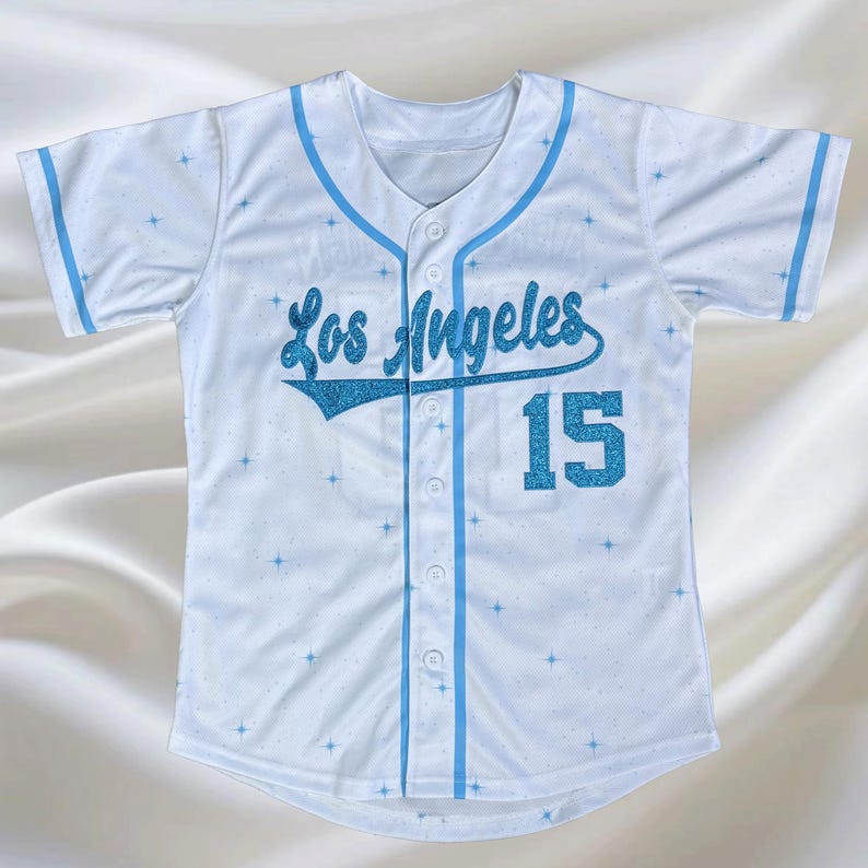 May include: White baseball jersey with light blue accents and the text "Los Angeles" in glittery blue script. The number "15" is also in glittery blue. The jersey has a button-up front and a V-neck.