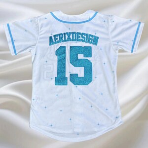 May include: White baseball jersey with light blue trim and accents. The jersey features the text "AERIXDESIGN" above the number "15" in a glittery blue. Small blue star patterns are scattered across the jersey.
