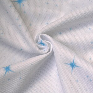May include: White fabric with a subtle texture, adorned with a pattern of small and large blue starbursts. The fabric is gathered, creating soft folds and shadows, highlighting the star design. The material appears to be lightweight.
