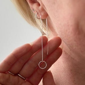 May include: A pair of silver threader earrings. One earring features a delicate chain with a small, open circle at the end. The other earring is a simple, straight silver bar. The earrings are shown in a close-up view.