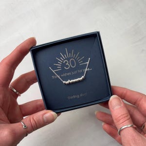 May include: A silver necklace with a series of small beads on a delicate chain, presented in a dark blue square box. The box insert features a sunburst graphic, the number 30, and the text Thirty wishes just for you... Sterling silver.