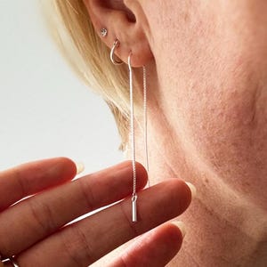 May include: Silver threader earrings. The earrings have a fine chain that dangles from the ear, with a small, cylindrical bar at the end. The image is a close-up, showing the minimalist design and the reflective quality of the silver. The earrings are a simple, elegant design.