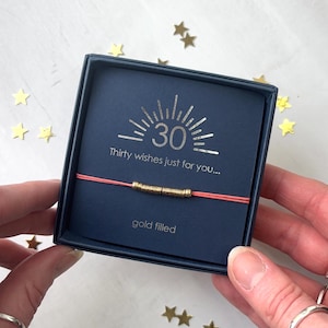 May include: A gold-filled bracelet with a coral cord is displayed in a navy blue gift box. The box is printed with the number "30", the text "Thirty wishes just for you..." and "gold filled". Gold star confetti is scattered around the box.