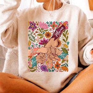 May include: Cream-colored sweatshirt featuring a floral design with the word "Mama" and several hands intertwined. The design includes colorful flowers and the word "Name" on the sleeves. A cozy and stylish top.