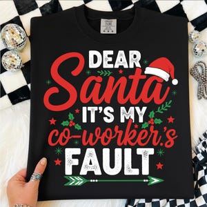 May include: Black crewneck sweatshirt with the text "DEAR Santa IT'S MY co-worker's FAULT" in red and white. The design includes a Santa hat, holly, and stars. A festive holiday-themed apparel item.