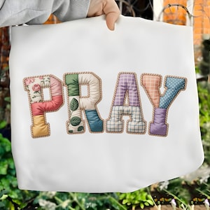 May include: White tote bag featuring the word "PRAY" in colorful, patchwork-style letters. Each letter is a different pattern, including floral, plaid, and polka dots, outlined with a rope-like trim.