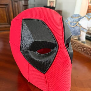 May include: A red and black Deadpool mask. The mask is primarily red with a textured fabric and features black eye and mouth details. The mask is designed to cover the entire head.