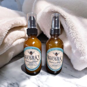 May include: Two amber glass bottles with black pump tops and labels that say "BeesSilky Magical Relief Plus+ Artisan's Apothecary All Natural Magnesium Gel". The bottles are sitting on a white marble surface.