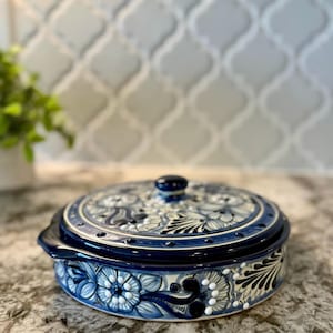 May include: A blue and white ceramic casserole dish with a lid. The dish is decorated with floral designs and white dots. The dish is on a gray countertop.