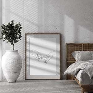 May include: A black and white line drawing of two hands intertwined, printed on a white background and framed in a brown frame. The print is hanging on a white wall in a room with a bed and a plant.