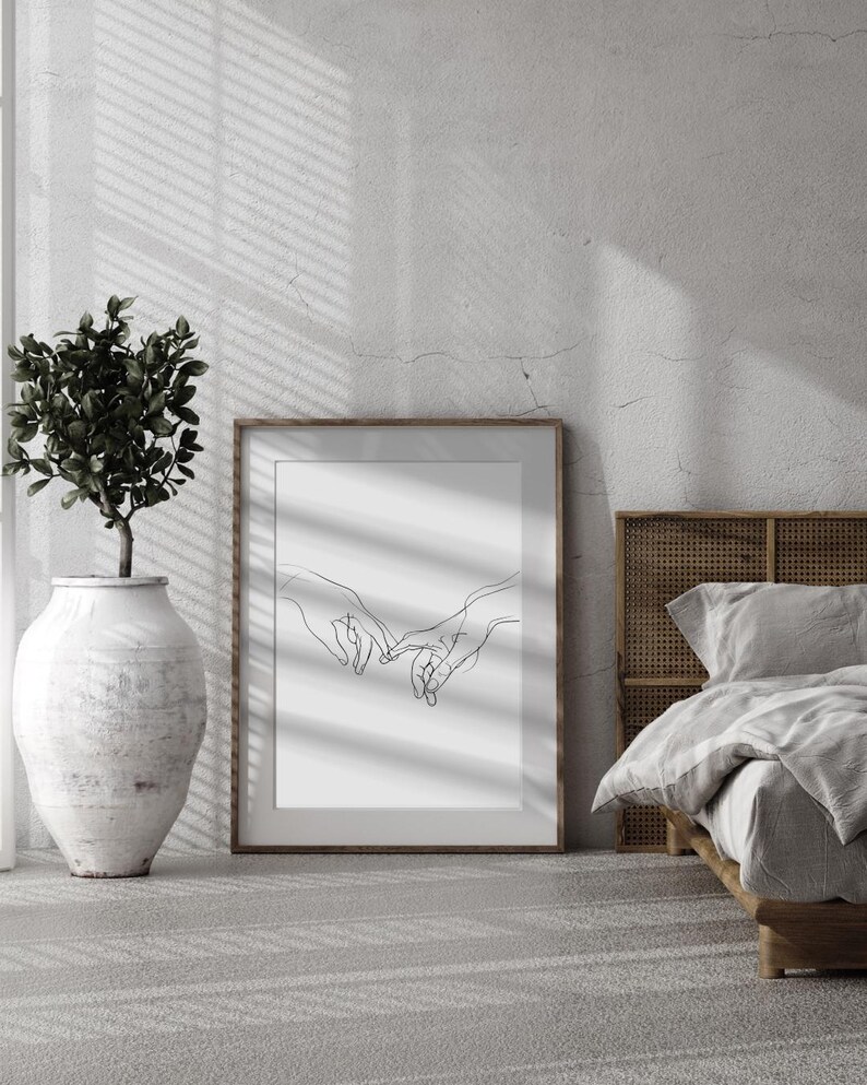 May include: A black and white line drawing of two hands intertwined, printed on a white background and framed in a brown frame. The print is hanging on a white wall in a room with a bed and a plant.