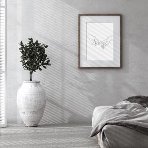May include: A minimalist bedroom with a white wall, a wooden bed frame, and a framed black and white line drawing of two hands intertwined. A potted plant sits on the floor next to the wall.
