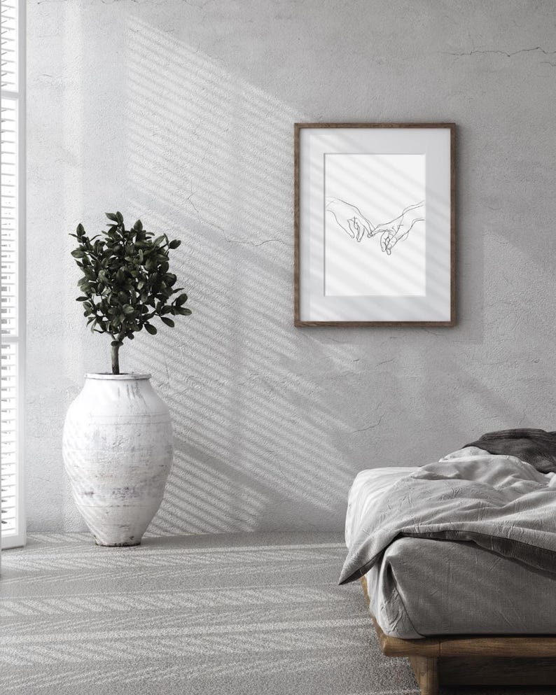 May include: A minimalist bedroom with a white wall, a wooden bed frame, and a framed black and white line drawing of two hands intertwined. A potted plant sits on the floor next to the wall.