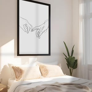 May include: Black and white line art print of two hands intertwined, framed in a black frame and hanging on a white wall above a bed with white bedding and beige throw pillows.