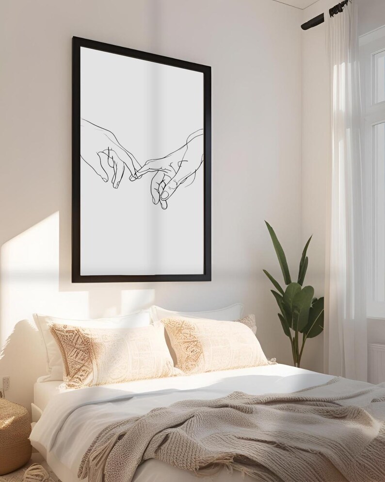 May include: Black and white line art print of two hands intertwined, framed in a black frame and hanging on a white wall above a bed with white bedding and beige throw pillows.