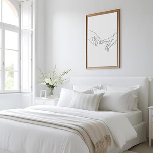 May include: A white bed with a white duvet cover and a beige throw blanket. The bed has a white headboard and a white pillow with a gray stripe pattern. There is a framed print of a line drawing of two hands reaching towards each other on the wall behind the bed.