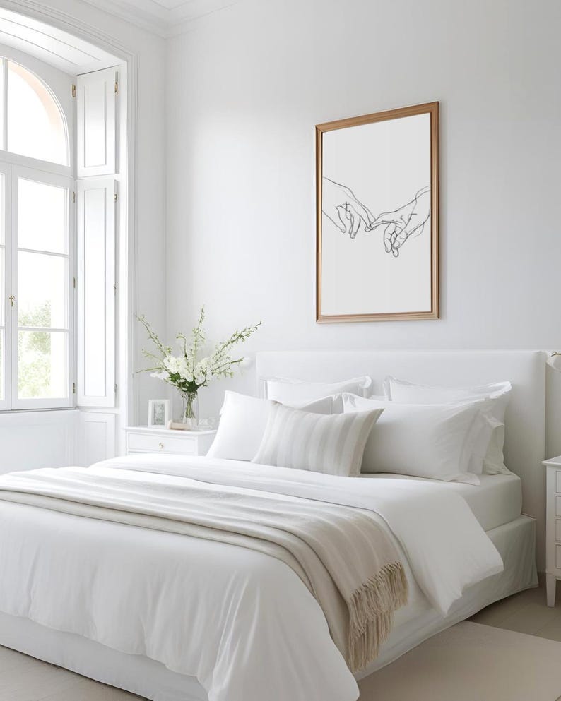 May include: A white bed with a white duvet cover and a beige throw blanket. The bed has a white headboard and a white pillow with a gray stripe pattern. There is a framed print of a line drawing of two hands reaching towards each other on the wall behind the bed.