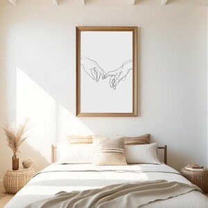 May include: A black and white line drawing of two hands intertwined, framed in a light brown frame, hanging on a white wall above a bed with white bedding and beige throw pillows.