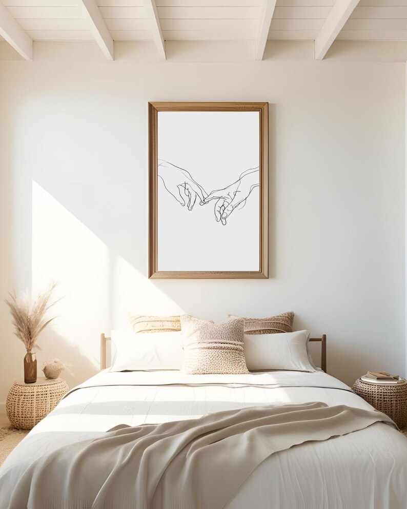 May include: A black and white line drawing of two hands intertwined, framed in a light brown frame, hanging on a white wall above a bed with white bedding and beige throw pillows.