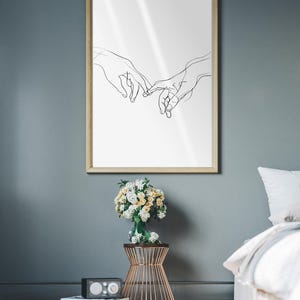 May include: A black and white line drawing of two hands reaching towards each other, framed in a light wood frame and hanging on a gray wall. A bouquet of white and yellow flowers is on a gold metal stand in front of the frame. A white bed with a white and gray blanket is in the background.