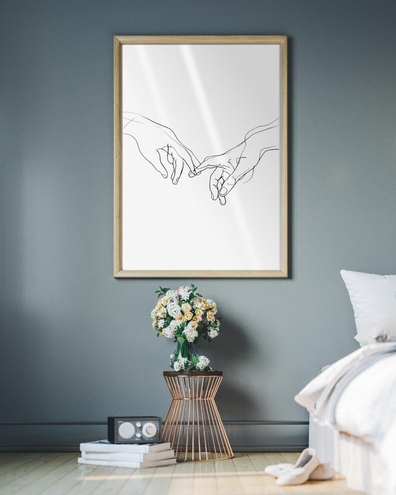 May include: A black and white line drawing of two hands reaching towards each other, framed in a light wood frame and hanging on a gray wall. A bouquet of white and yellow flowers is on a gold metal stand in front of the frame. A white bed with a white and gray blanket is in the background.