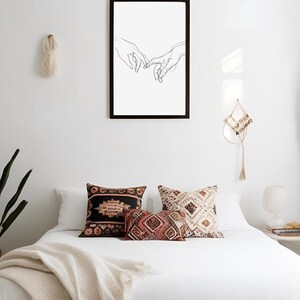 May include: A black framed print of a line drawing of two hands reaching towards each other. The print is hanging on a white wall above a white bed with a white blanket and three patterned pillows.