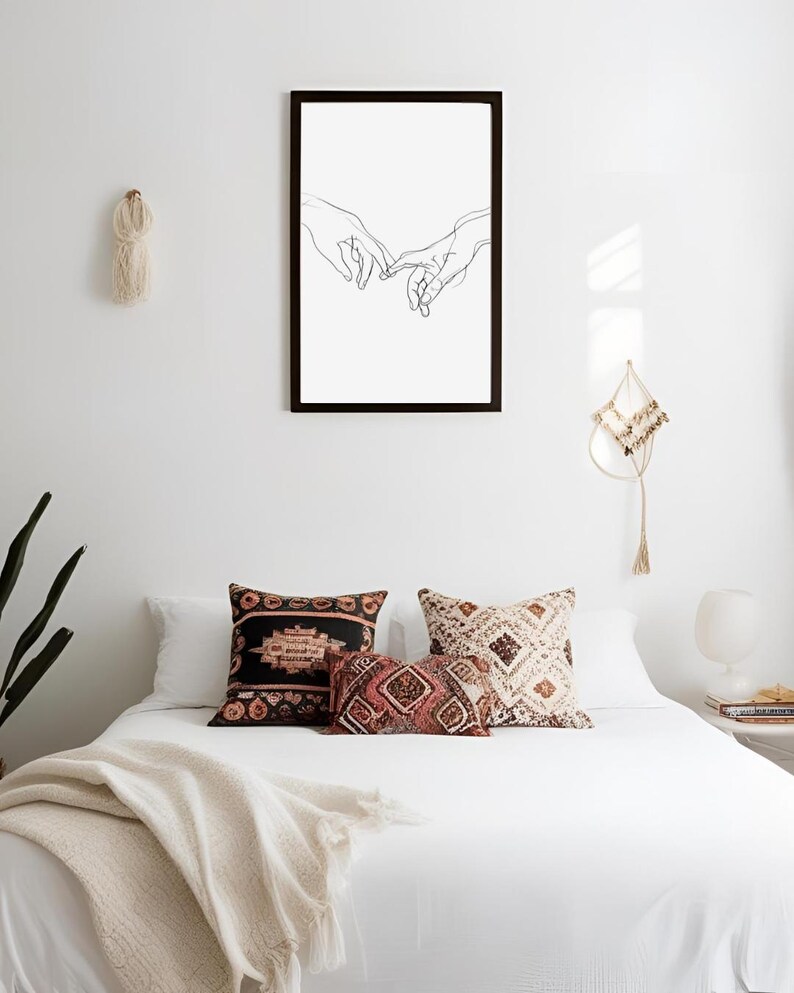 May include: A black framed print of a line drawing of two hands reaching towards each other. The print is hanging on a white wall above a white bed with a white blanket and three patterned pillows.