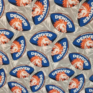 May include: A gray fabric featuring a repeating pattern of blue American footballs. Each football displays the word DENVER in white capital letters above an orange horse head logo. The background is decorated with faint, light gray mountain outlines.