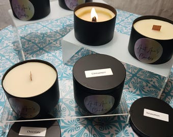 Puede incluir: Una colección de velas de lata negras con etiquetas y tapas blancas. Las velas tienen cera de color claro y están etiquetadas con aromas como "Canela", "Chocolate" y "Relaxación". Una vela está encendida, mostrando una llama.