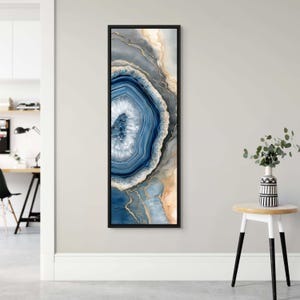 Puede incluir: Una pieza de arte mural rectangular vertical que presenta un patrón abstracto de piedra ágata en tonos azul marino profundo, gris suave y beige cálido con acentos de vetas doradas metálicas. La obra está enmarcada en un marco negro fino, mostrando capas concéntricas y un centro cristalino que imita la apariencia natural de una rodaja de piedra preciosa pulida.