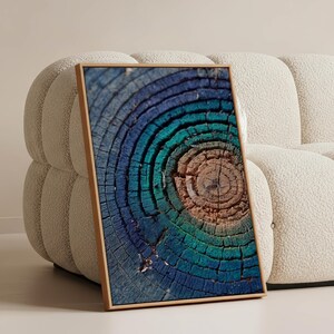 Op de afbeelding: Een ingelijst kunstwerk met een close-up van de dwarsdoorsnede van een boomstam. De houtnerf toont concentrische ringen in blauwe, turquoise en bruine tinten. Het kunstwerk wordt gepresenteerd tegen een neutraal getinte, getextureerde bank.