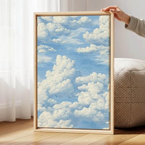 May include: A framed artwork featuring a blue sky filled with fluffy white clouds. The artwork is in a light-colored wooden frame. The image is held by a person's hand, suggesting it's a piece of home decor.
