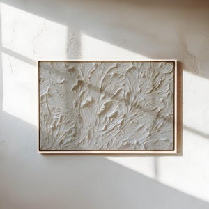 May include: A rectangular, textured, off-white abstract art piece in a light wood frame. The artwork features a raised, flowing design, resembling a stylized natural form. The piece is mounted on a white wall, with shadows from sunlight.