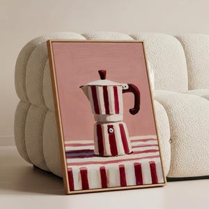 May include: A framed painting of a red and white striped stovetop espresso maker. The painting has a pink background and is leaning against a cream-colored sofa. The espresso maker has a dark brown handle and top.