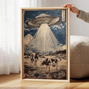 May include: A framed print depicting a UFO beaming light onto two cows in a field, with a mountain in the background. The artwork is in a woodblock print style, with blue and beige tones. The print is held by a person.