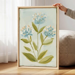 Könnte beinhalten: Gerahmtes Gemälde mit blauen und weißen Hortensienblüten und grünen Blättern vor hellblauem Hintergrund. Das Kunstwerk befindet sich in einem Holzrahmen und wird von einer Person gehalten.