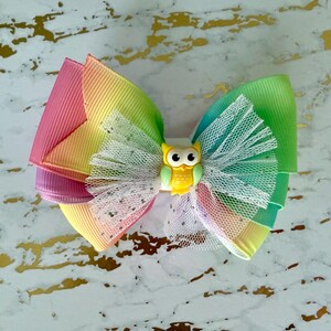 May include: A colorful hair bow featuring layers of rainbow-colored ribbon and white tulle. A small yellow owl charm is centered on the bow. The bow is a vibrant accessory.