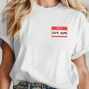 Puede incluir: Una camiseta blanca de manga corta con un gráfico de etiqueta de nombre rectangular en rojo y blanco en el pecho izquierdo. La etiqueta incluye el texto HELLO my name is en blanco y negro, con las palabras YOUR NAME escritas en estilo marcador.