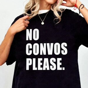 May include: Black t-shirt with the text "NO CONVOS PLEASE" in large white capital letters. The shirt is a casual style, suitable for everyday wear. The model is wearing a gold necklace.