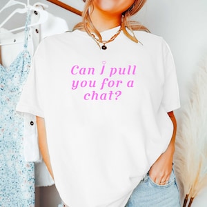以下が含まれることがあります： 「Can I pull you for a chat?」というピンクの文字と、その上に小さなピンクのハートのアイコンがプリントされた白い半袖Tシャツ。クルーネックでゆったりとしたシルエットのTシャツに、ブルーのデニムジーンズとゴールドのネックレスを合わせています。