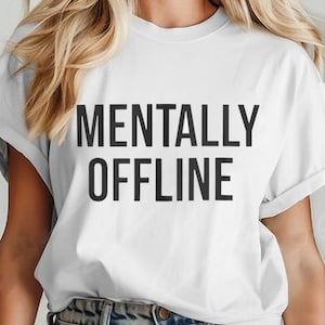 Puede incluir: Camiseta blanca con las palabras "MENTALLY OFFLINE" impresas en negrita y letras mayúsculas negras. La camiseta tiene mangas cortas y cuello redondo. La modelo lleva vaqueros azules.