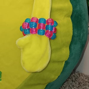 May include: A colorful bracelet made of translucent pink and blue beads is displayed on a yellow plush toy. The bracelet is made of square beads and is worn on the toy's arm. The toy is a light green and yellow color.
