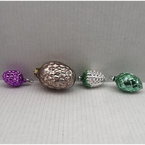May include: Four vintage glass Christmas ornaments in a row. The ornaments are in the shape of fruits and vegetables. The first ornament is purple, the second is silver, the third is green and silver, and the fourth is green.